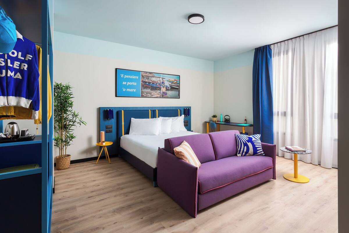 Ett hotellrum med en säng, en lila soffa, ett litet bord, en krukväxt och en tavla på väggen.