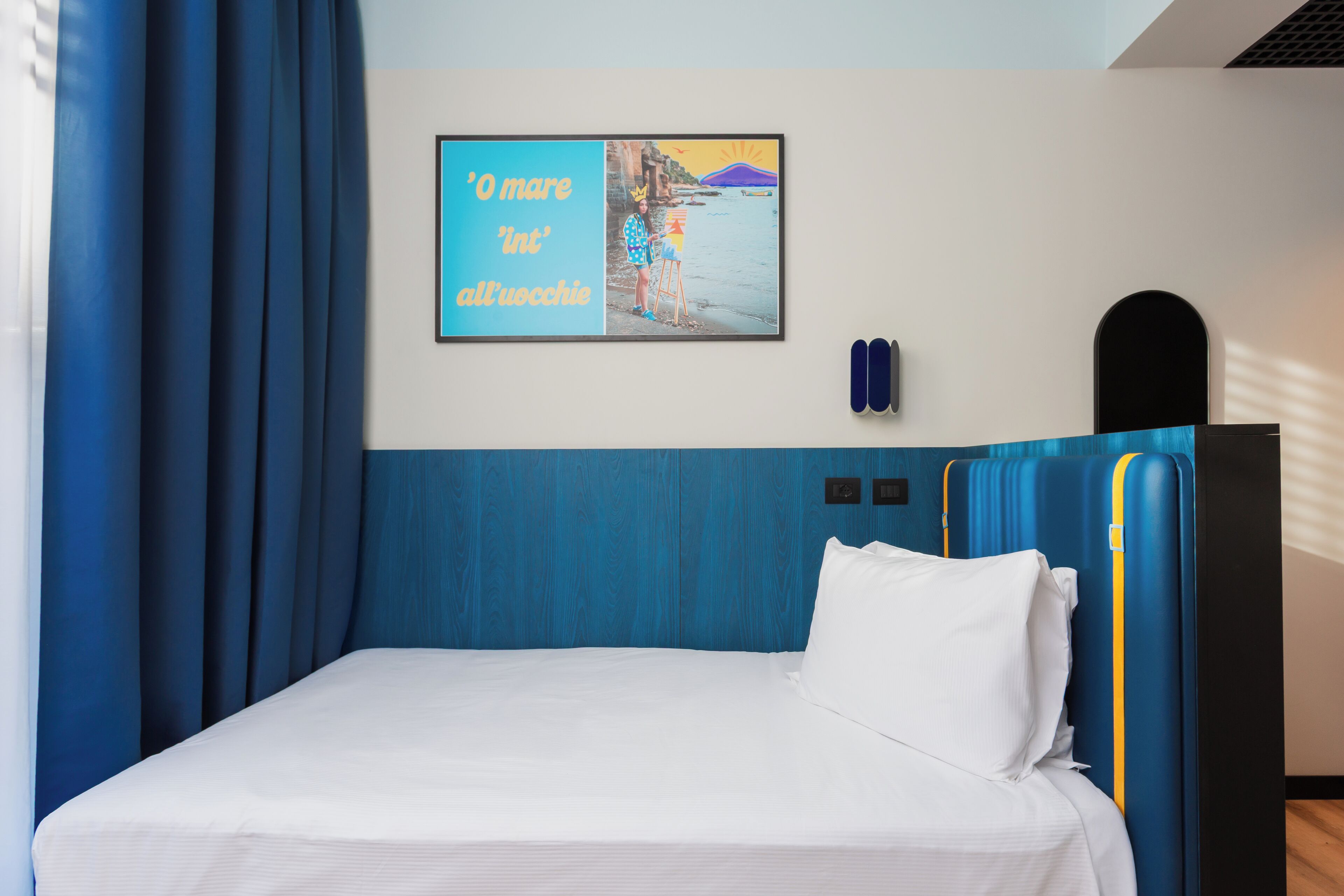 A hotel room with a bed, a wall-mounted picture, and a window with curtains.