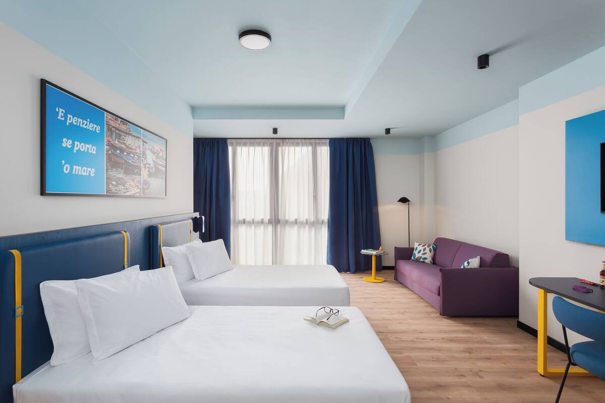 Ett hotellrum med två sängar, en lila soffa, ett litet bord och ett stort fönster med gardiner.