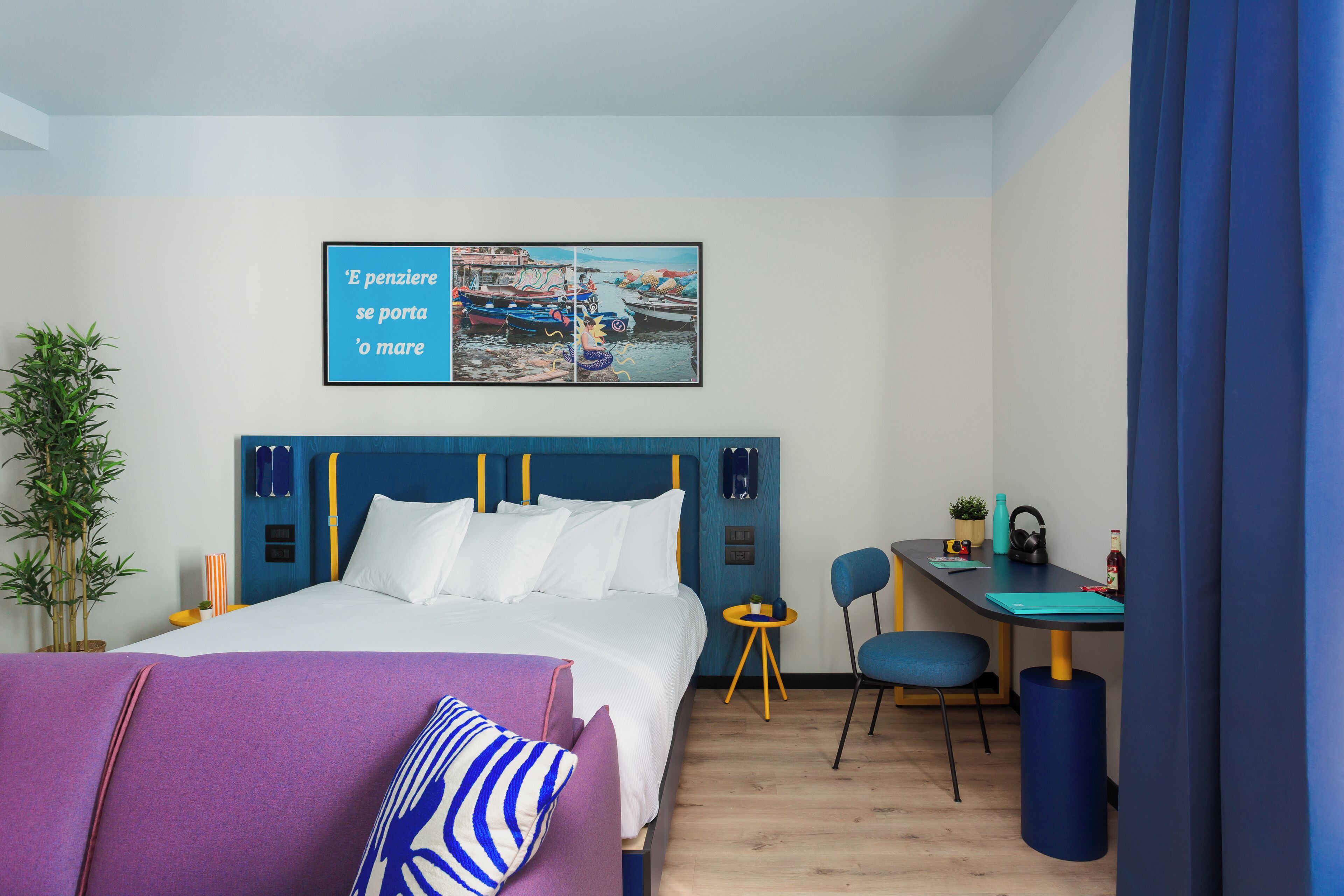 A hotel room with a bed, a desk, a chair, a potted plant, and a wall art piece.