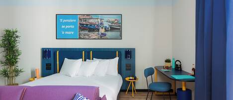 A hotel room with a bed, a desk, a chair, a potted plant, and a wall art piece.