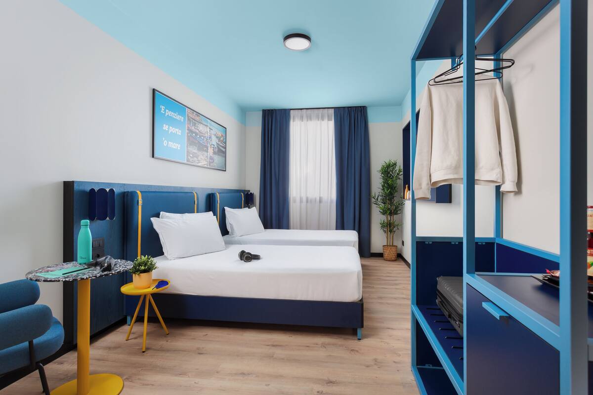Ett modernt hotellrum med en säng, ett skrivbord och en stol. Det finns ett fönster med gardiner, en växt på skrivbordet och en vägghylla med ett handduk.