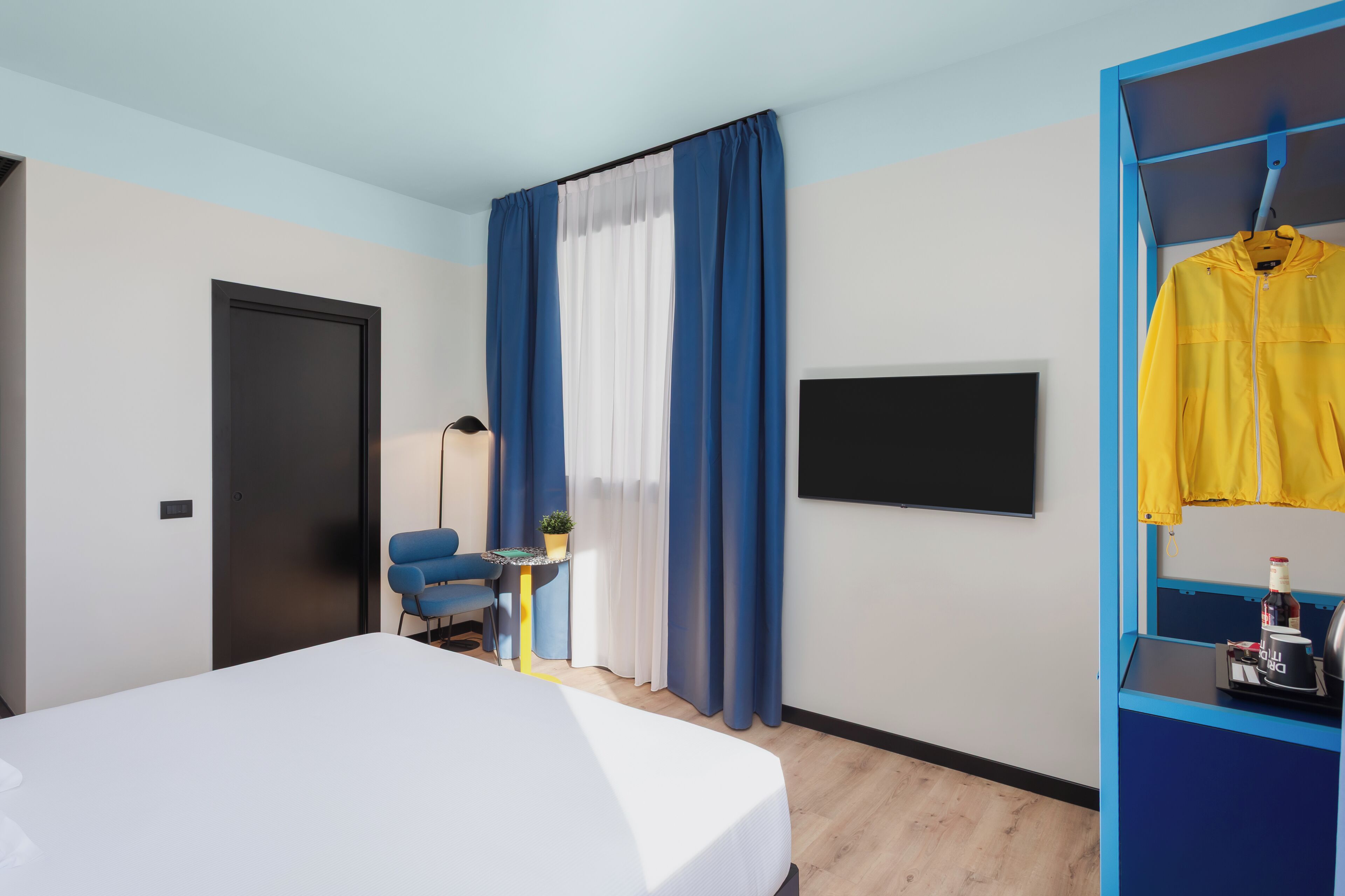 A modern hotel room with a bed, a desk, a chair, a TV, and a wardrobe with a yellow jacket hanging.