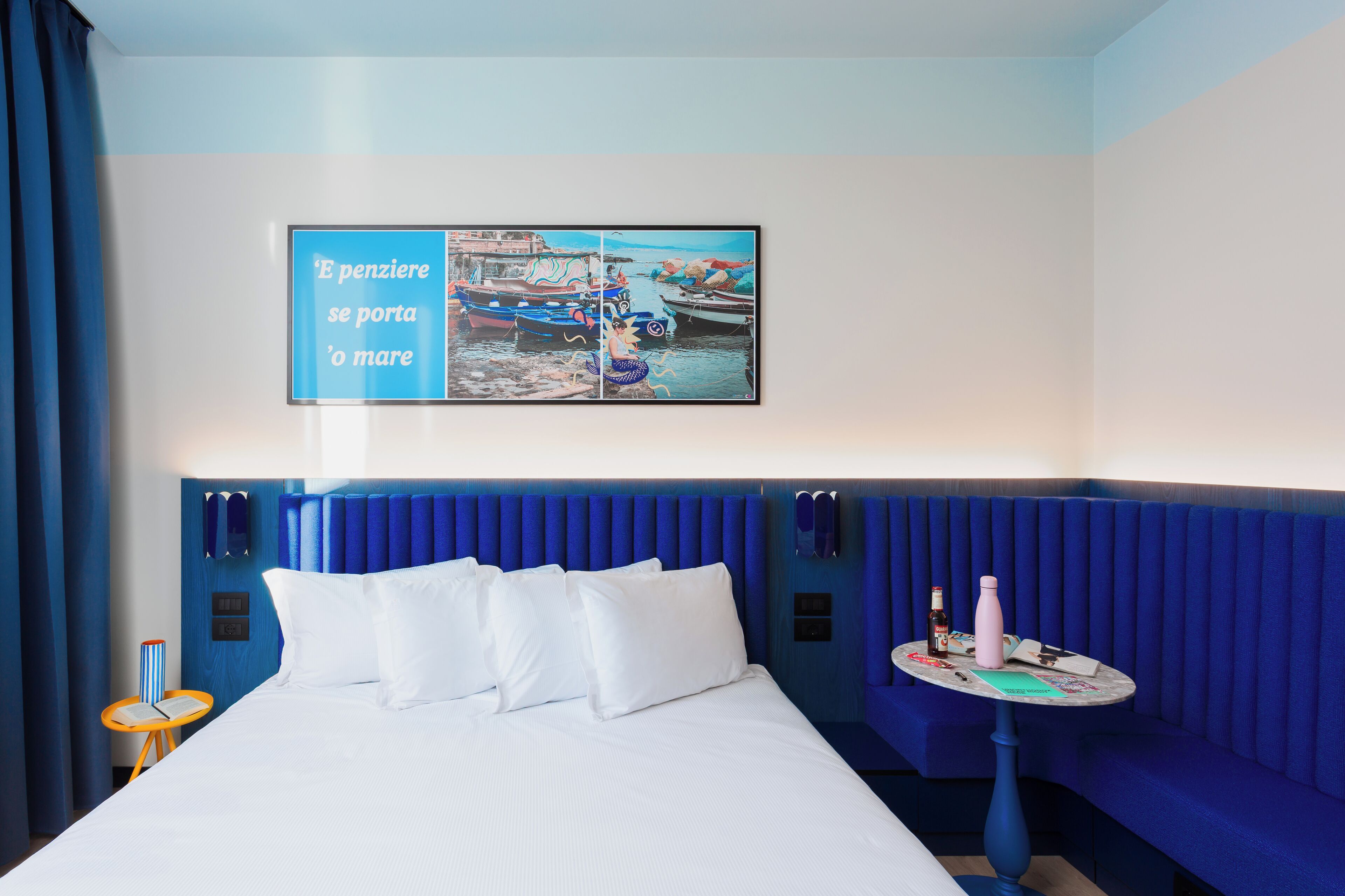 A hotel room with a bed, a blue headboard, a small table with a bottle and a book, and a wall with a framed picture.