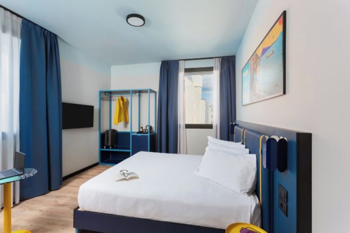Une chambre d’hôtel avec un lit, un bureau avec un ordinateur portable, un porte-manteau et un tableau au mur.