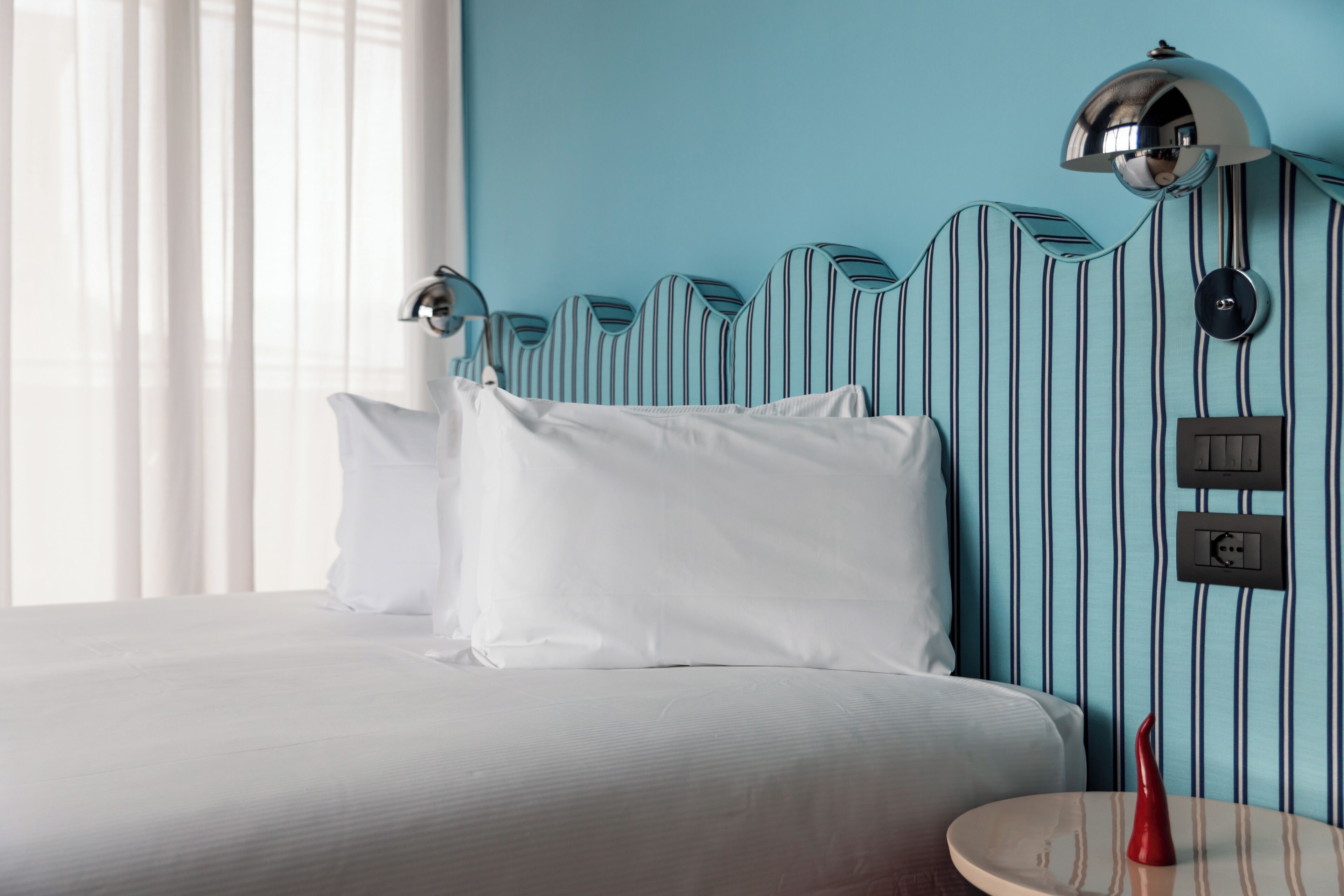 A hotel room with a bed, white pillows, a round table with a red vase, and a striped wall with a unique headboard design.