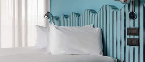 A hotel room with a bed, white pillows, a round table with a red vase, and a striped wall with a unique headboard design.