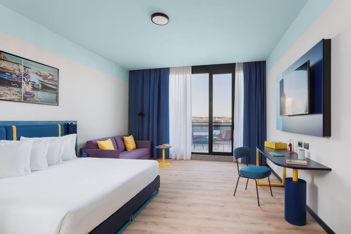 Ett modernt hotellrum med en stor säng, ett skrivbord, en stol och utsikt över staden.