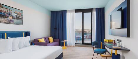 A modern hotel room with a large bed, a desk, and a view of the city through the window.