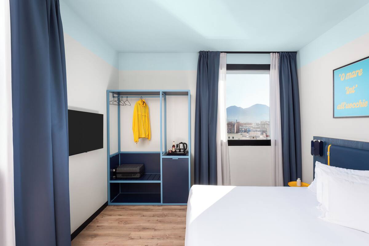 Ett hotellrum med en säng, en garderob, ett fönster med utsikt och en vägg med en affisch.