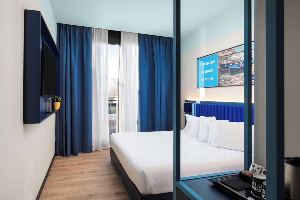 Ett hotellrum med en säng, en tv som är monterad på väggen, ett fönster med gardiner och utsikt över båtar.