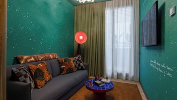 A room with teal walls, a dark sofa with patterned cushions, a round coffee table with books and a red object, and a large window with sheer curtains.