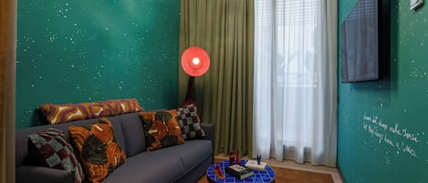 A room with teal walls, a dark sofa with patterned cushions, a round coffee table with books and a red object, and a large window with sheer curtains.