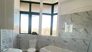 Een moderne badkamer met toilet, wastafel en bad. Twee ramen met groene kaders, en de muren zijn betegeld met een marmer dessin.