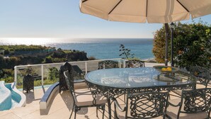 Ein Tisch mit Glasplatte, Stühle und Sonnenschirm mit Blick auf einen Pool und das Meer.
