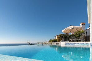 Une piscine d’un bleu éclatant avec vue sur la mer, un bâtiment blanc surmonté d’une tour et un espace détente agrémenté de parasols.