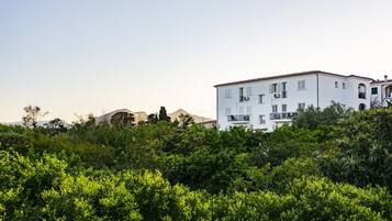 A white building with balconies, surrounded by dense greenery and mountains in the background.