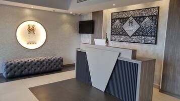 Un área de recepción moderna de hotel con un banco capitonado, un letrero circular iluminado, un televisor de pantalla plana y un elemento decorativo en la pared.