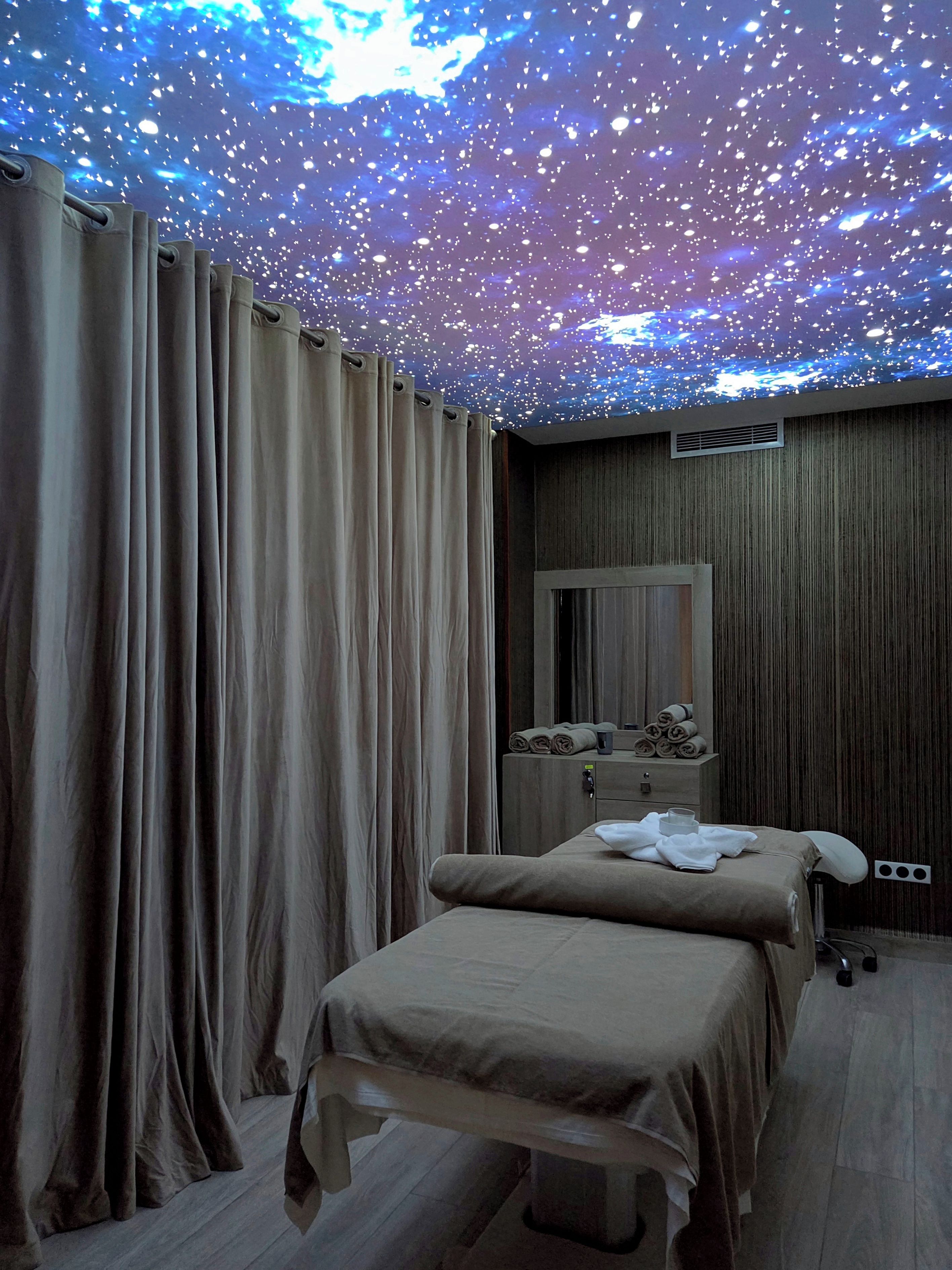 Habitación de hotel con cama, cortinas, tocador con espejo y techo con proyección de cielo estrellado.