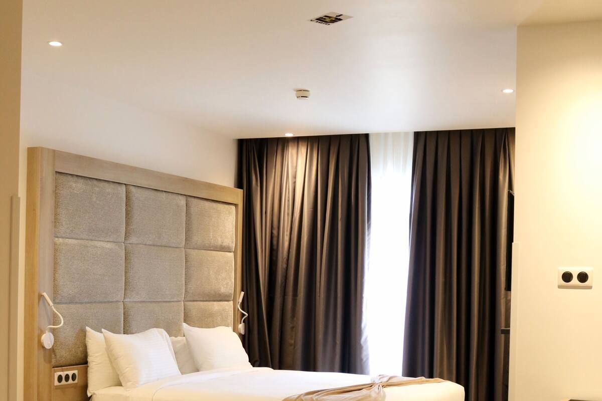 A modern hotel room with a large bed, a wooden nightstand, a desk, and a large window with curtains.