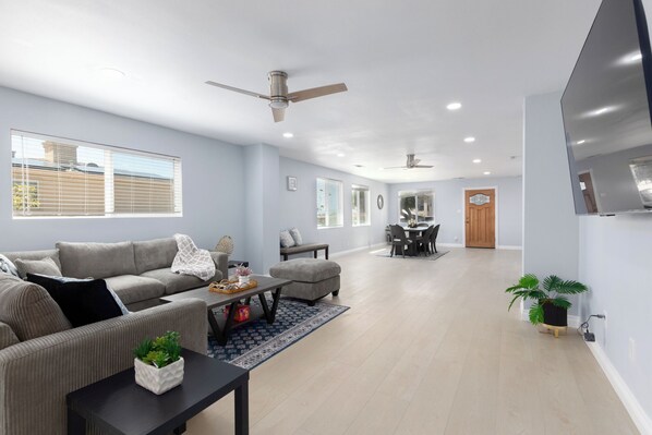 A spacious living room with a grey sofa, a coffee table with decorative items, a dining area with a table and chairs, and a TV mounted on the wall.
