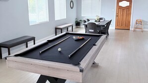 Una mesa de billar con bolas y tacos, un banco y una mesa de comedor con sillas en una habitación con paredes de colores claros y suelo de madera.