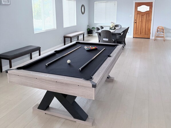 Une table de billard avec des billes et des queues, un banc et une table à manger avec des chaises, le tout dans une pièce aux murs de couleur claire et au plancher en bois.