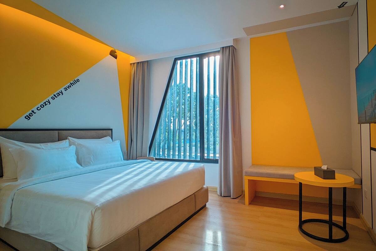 A modern hotel room with a bed, a small table, a TV, and a window with curtains.