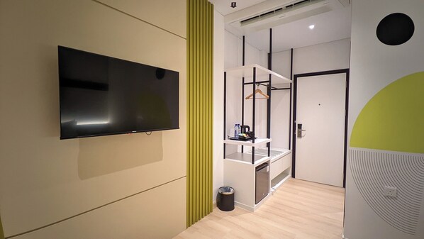 A modern hotel room with a flat-screen TV mounted on the wall, a small seating area with a table and chairs, and a coat rack.