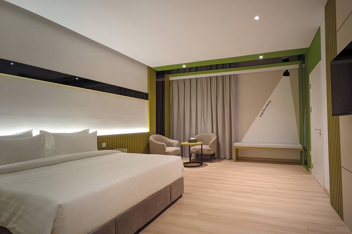 A modern hotel room with a large bed, two armchairs, a small table, and a TV mounted on the wall.