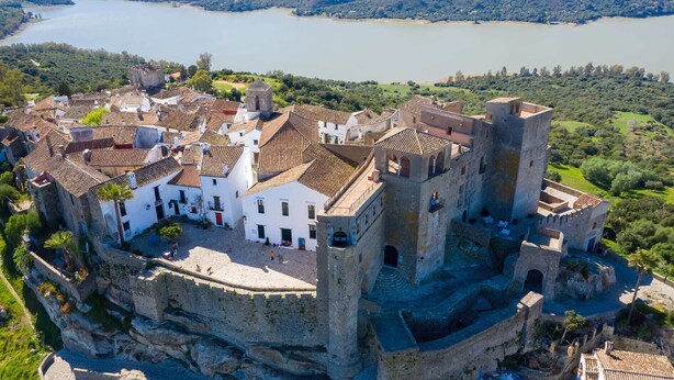 Un pueblo amurallado histórico con un castillo central, rodeado de edificios blancos con techos de tejas rojas, con vistas a un lago.