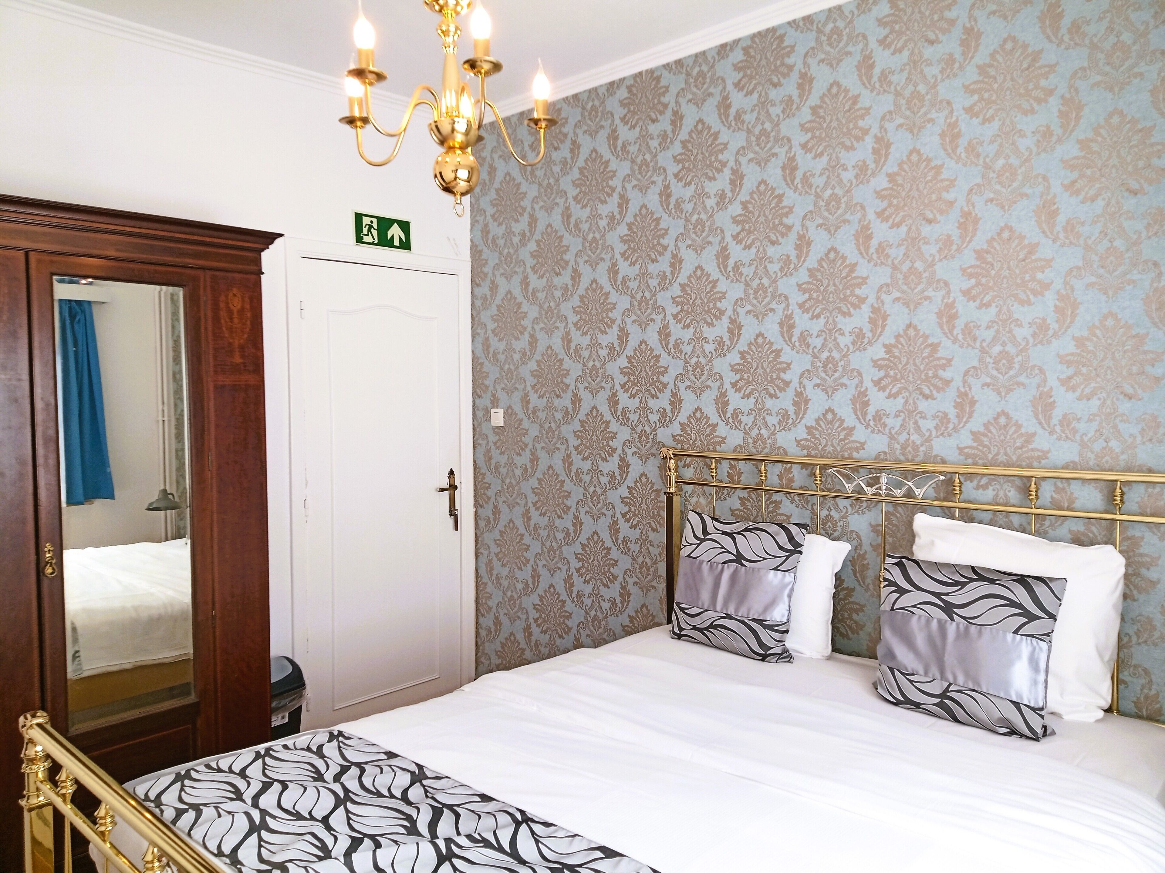 A hotel room with a bed, a chandelier, a door with an exit sign, and patterned wallpaper.