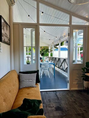 Ein Zimmer mit Holzboden, einem Sofa mit gemustertem Kissen und Blick auf eine Terrasse mit Tisch und Stühlen.