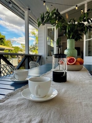 A table with a coffee pot, cups, and a bowl of fruit on a porch.