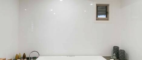 Un baño moderno con una bañera blanca grande, una ventana pequeña y diversos artículos de aseo en la encimera.
