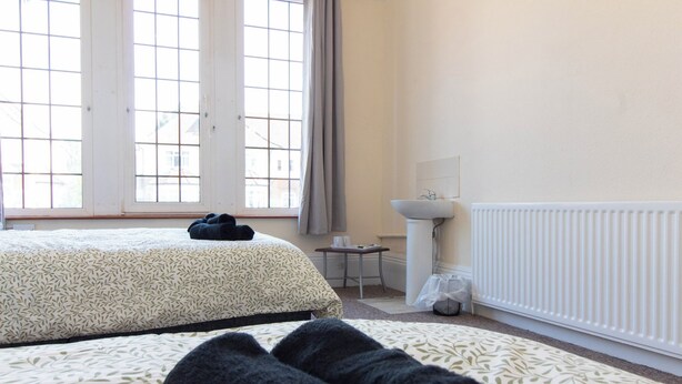 A hotel room with two beds, a window with curtains, a small table with a cup and a tissue, and a radiator. - Brookvale Guest House Room 2 (Southampton)