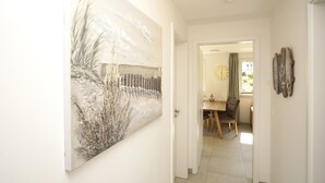 A hallway with a large abstract painting on the left, a dining area with a table and chairs in the background, and a clock on the wall. - Beachfront apartment with terrace, use of swimming pool and sauna - Rex Rugia H-1010 (Mönchgut OT Lobbe)