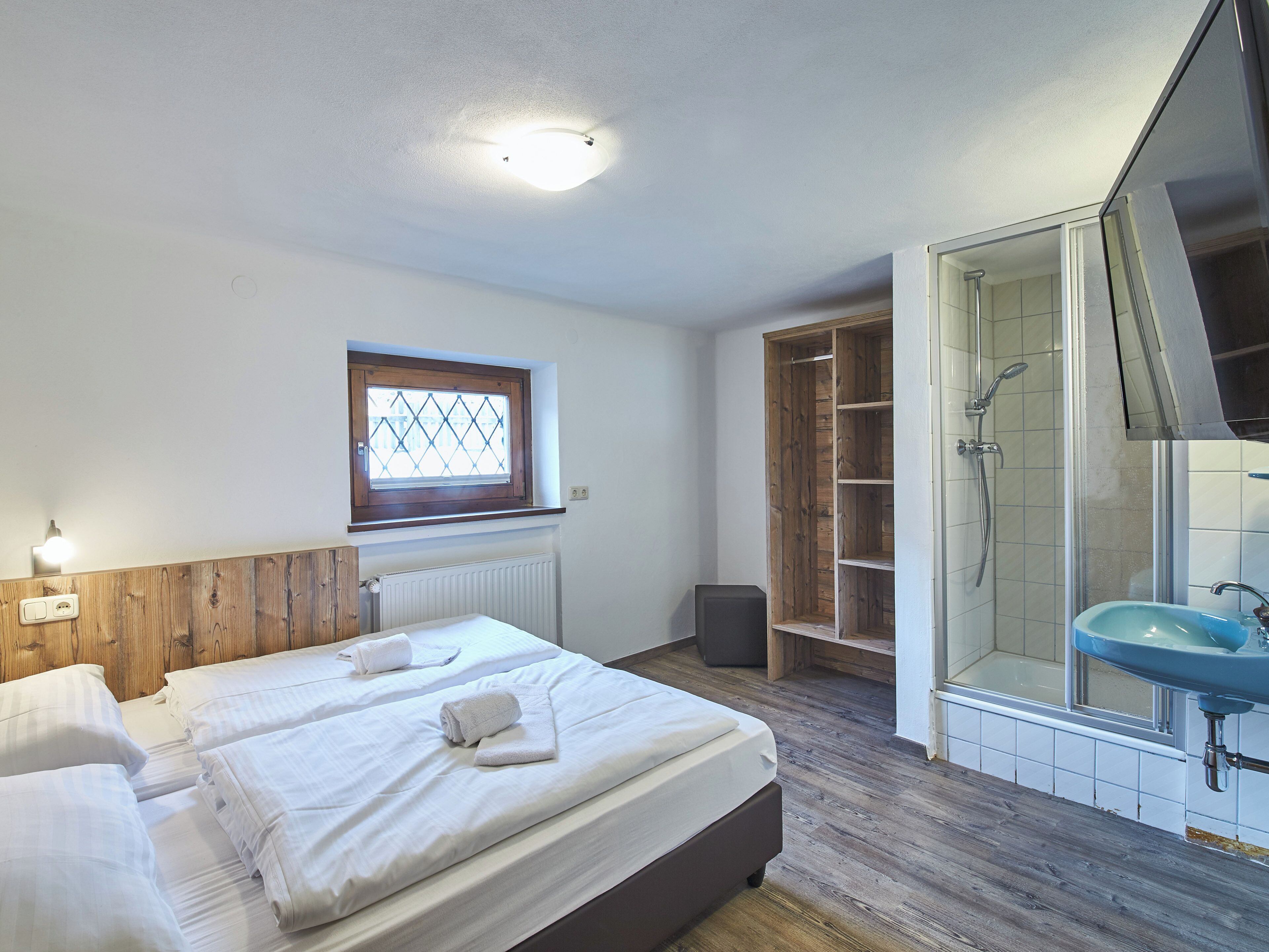 A bedroom with two beds, a window, a wooden wardrobe, a shower area, and a bathroom with a sink.