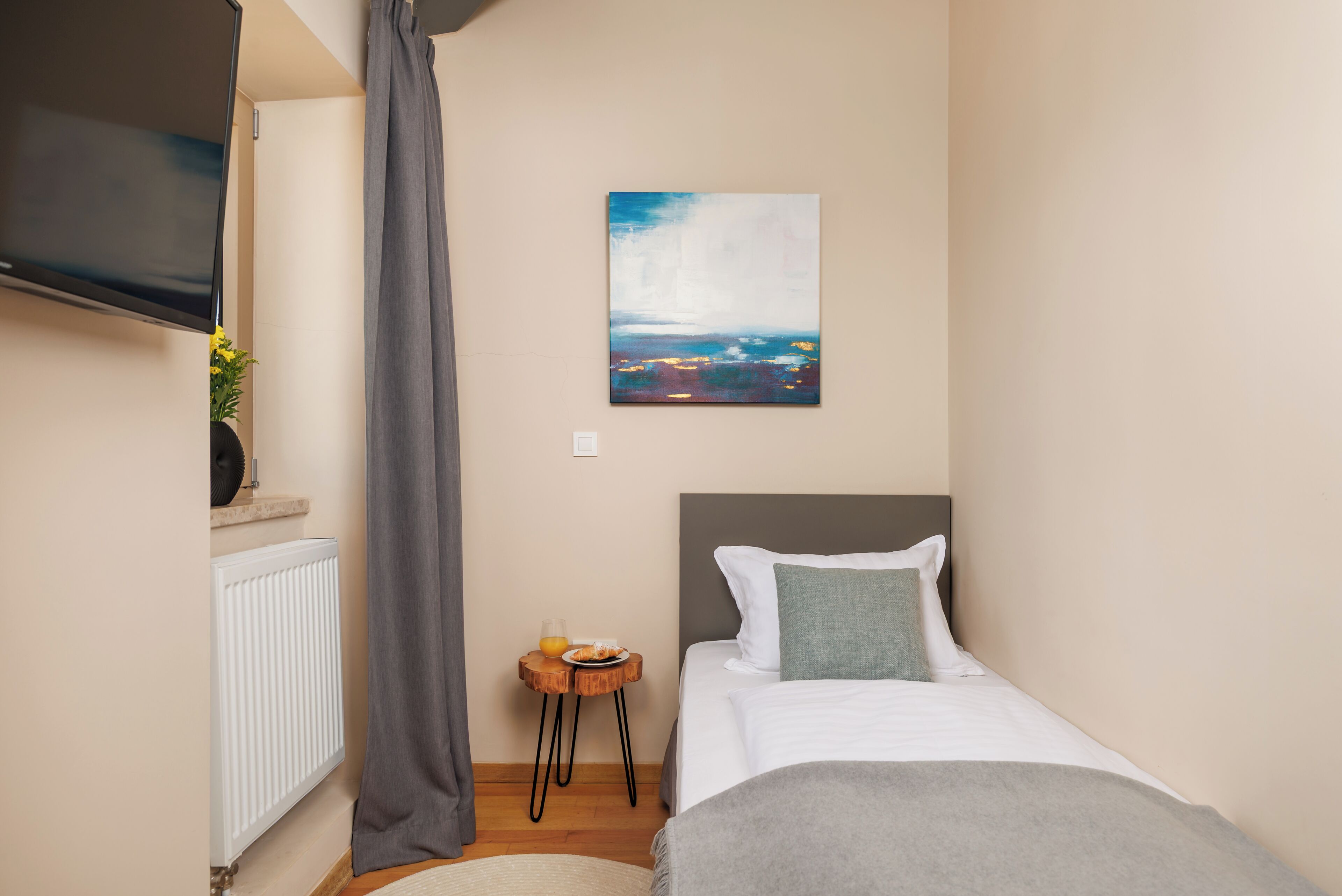 A bedroom with a bed, a TV mounted on the wall, a painting, a small table with a plate and a glass, and a radiator.