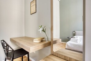 Een moderne kamer met een houten bureau, een stoel, een nachtkastje met een boek en een vaas, en een bed met witte lakens.
