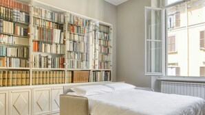 Une chambre avec une grande bibliothèque remplie de livres, un lit avec une literie blanche et une fenêtre donnant sur les bâtiments environnants.