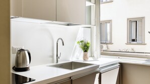 Une cuisine moderne équipée d’une bouilloire en acier inoxydable, d’un évier et d’une fenêtre donnant sur un immeuble.