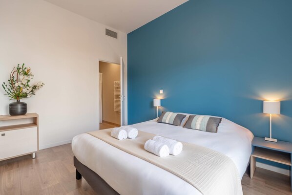 Una habitación de hotel moderna con una cama grande, dos lámparas de noche, una mesita de noche, una planta en maceta y vistas al pasillo.