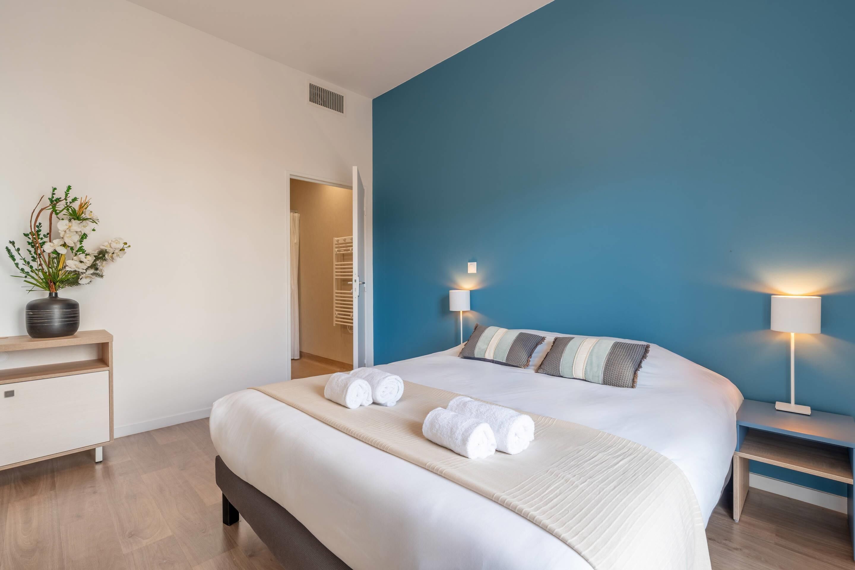 Una habitación de hotel moderna con una cama grande, dos lámparas de noche, una mesita de noche, una planta en maceta y vistas al pasillo.