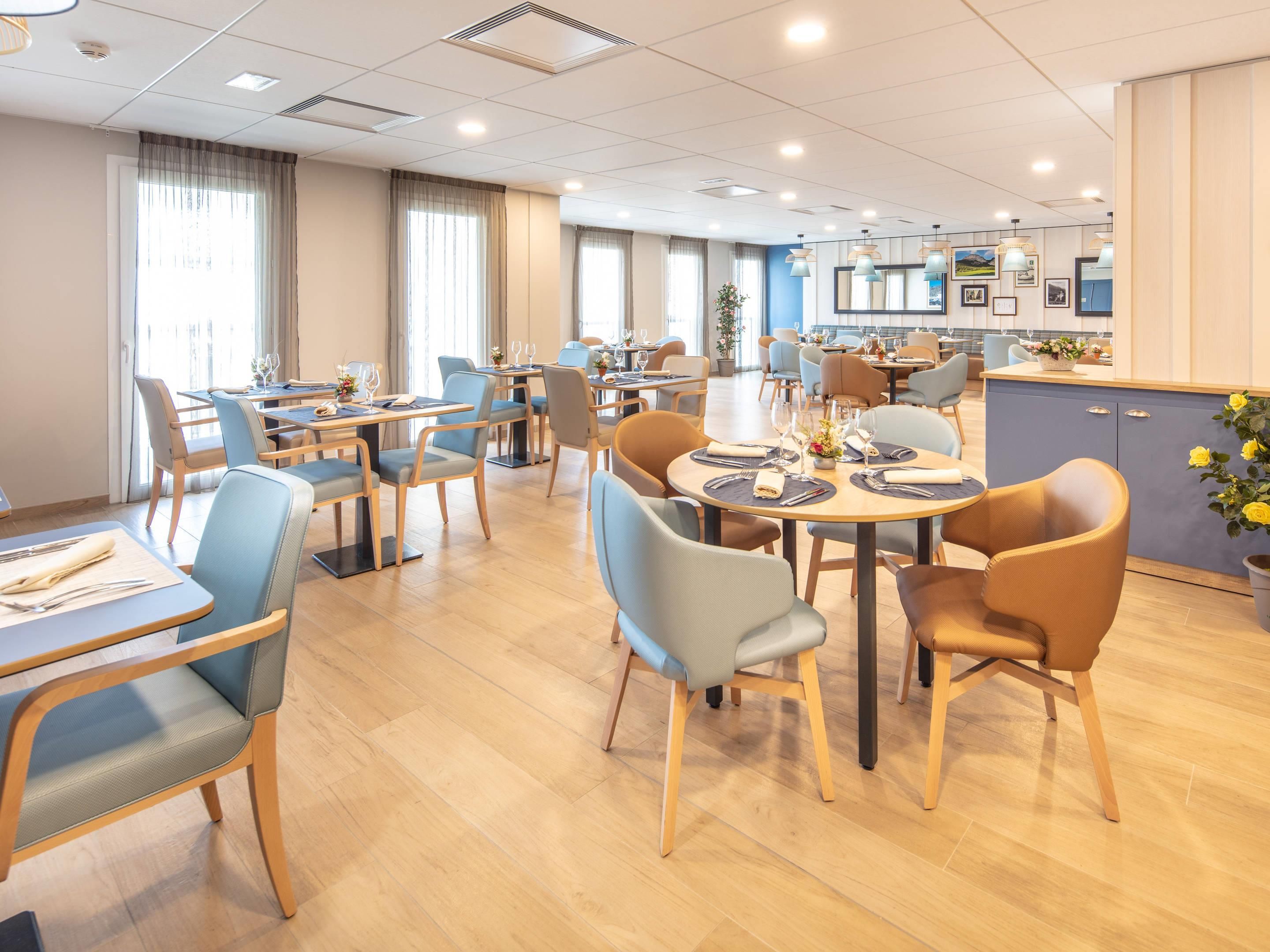 A hotel dining area with multiple tables and chairs, a kitchen in the background, and large windows allowing natural light.