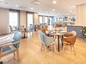 Een restaurantruimte in een hotel met meerdere tafels en stoelen, een keuken op de achtergrond en grote ramen voor natuurlijk lichtinval.