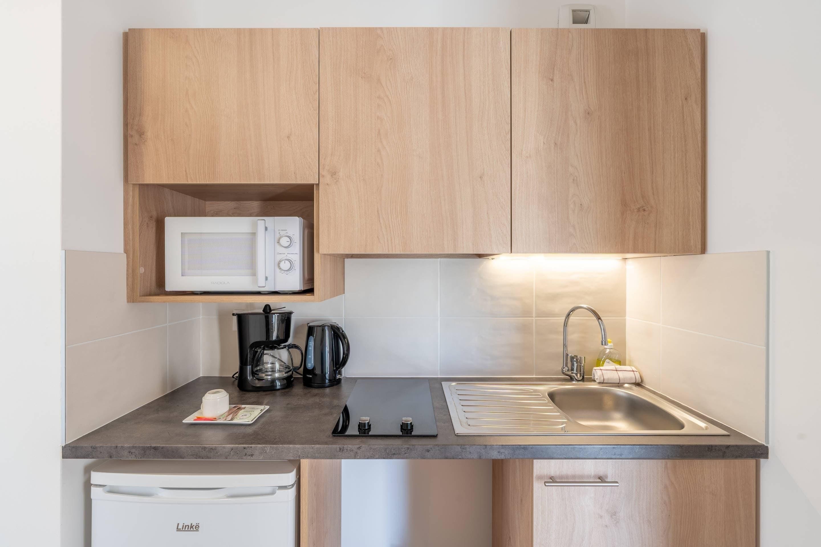 A compact kitchen with wooden cabinets, a microwave, a coffee maker, a kettle, a sink, and a dishwasher.
