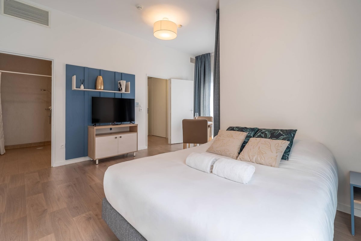 Une chambre d’hôtel moderne avec un grand lit, un téléviseur posé sur une commode bleue, un bureau et une chaise.