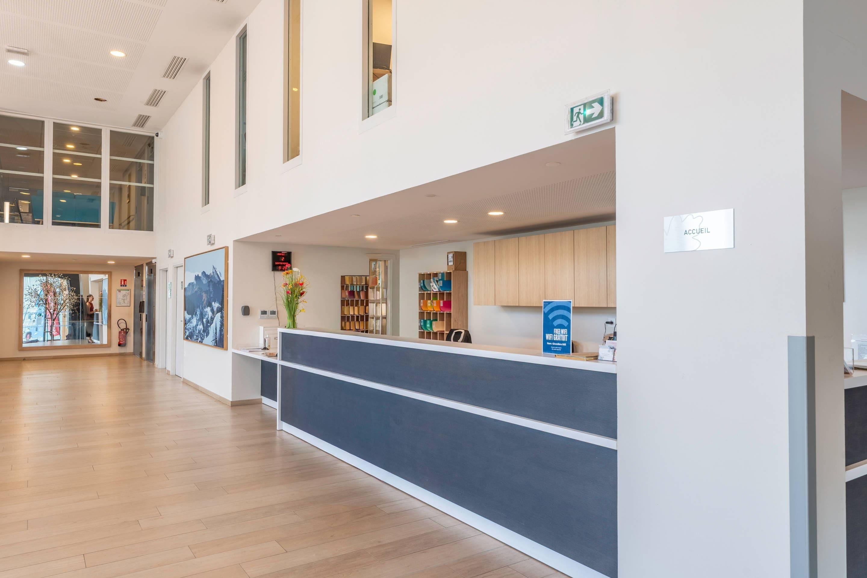 Un hall d’entrée spacieux avec un comptoir de réception, un plancher en bois et une vue sur l’intérieur grâce à de grandes fenêtres.
