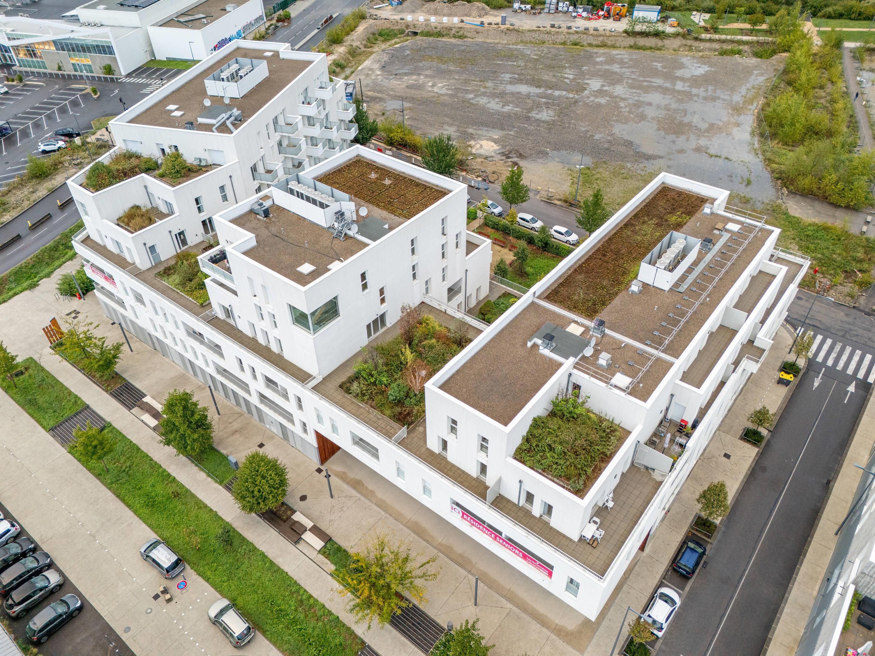 A modern multi-story residential building with rooftop gardens, a parking lot, and a road in the background.